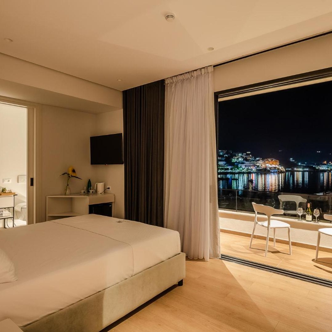 Hotel room with a bed facing open glass doors onto a balcony above the lit bay at Rea Boutique Hotel in Himarë