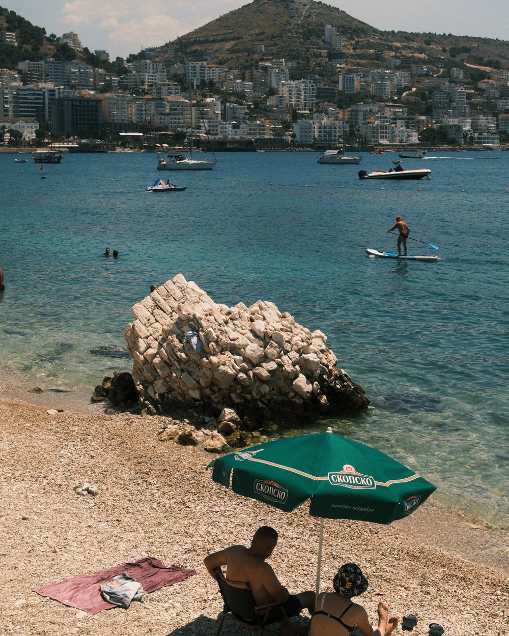 Paddleboarder and boats on clear turquoise water beside a large rock at Sarandë main beach, Albania