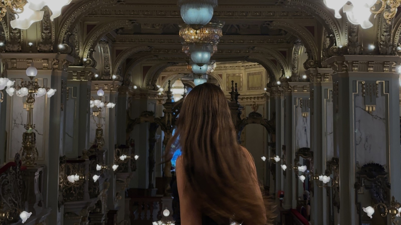 Woman standing in the gilded interior of New York Cafe in Budapest beneath the chandeliers
