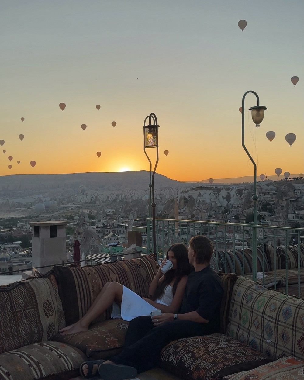 Couple sitting on cushioned rooftop seating at sunrise with hot air balloons rising over the valley