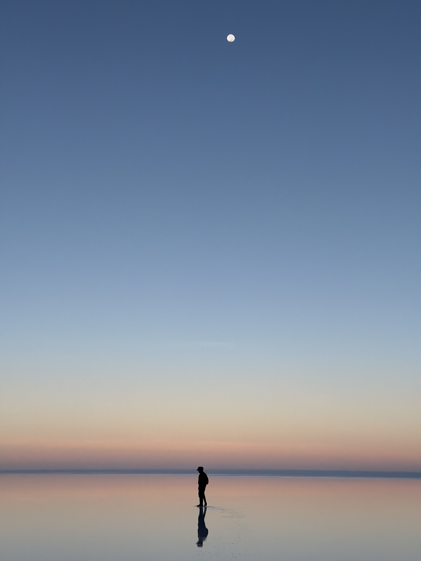 A traveller and their reflection beneath the moon on the Uyuni salt flats at dawn