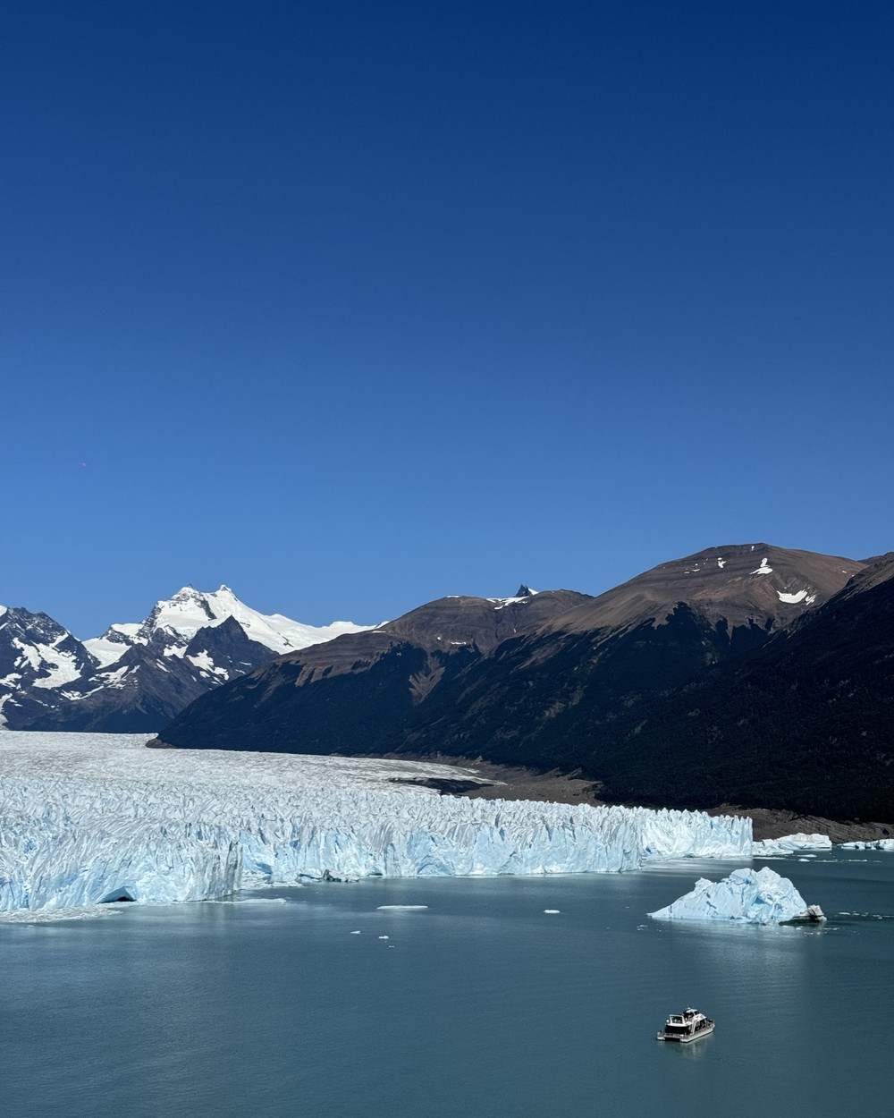 One small boat on the lake below the whole glacier front, with snowy peaks behind and an iceberg nearby