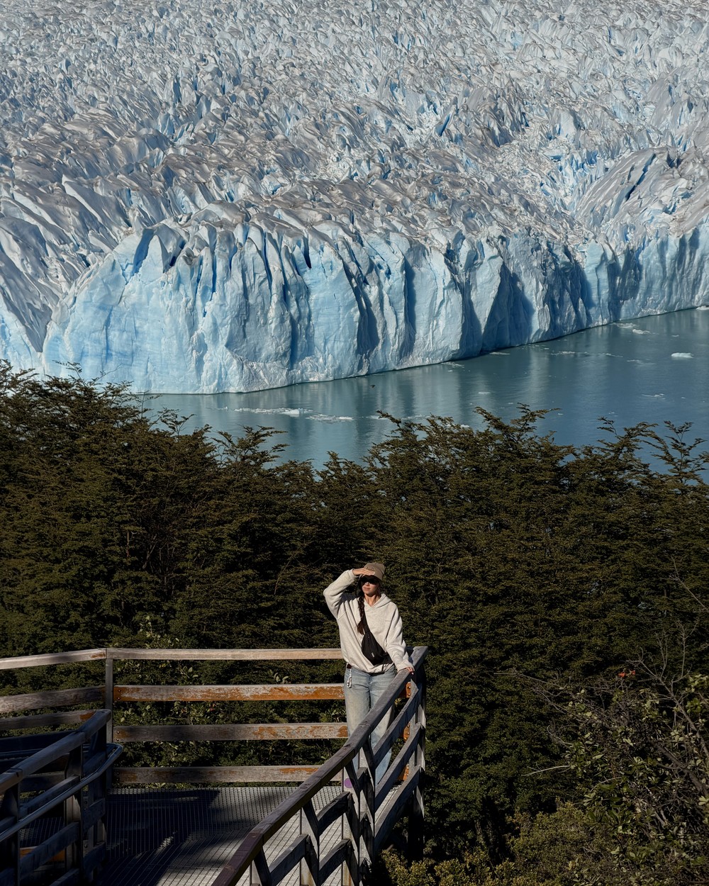 Ellen on a wooden walkway platform among the trees, with the Perito Moreno glacier face and the lake behind her