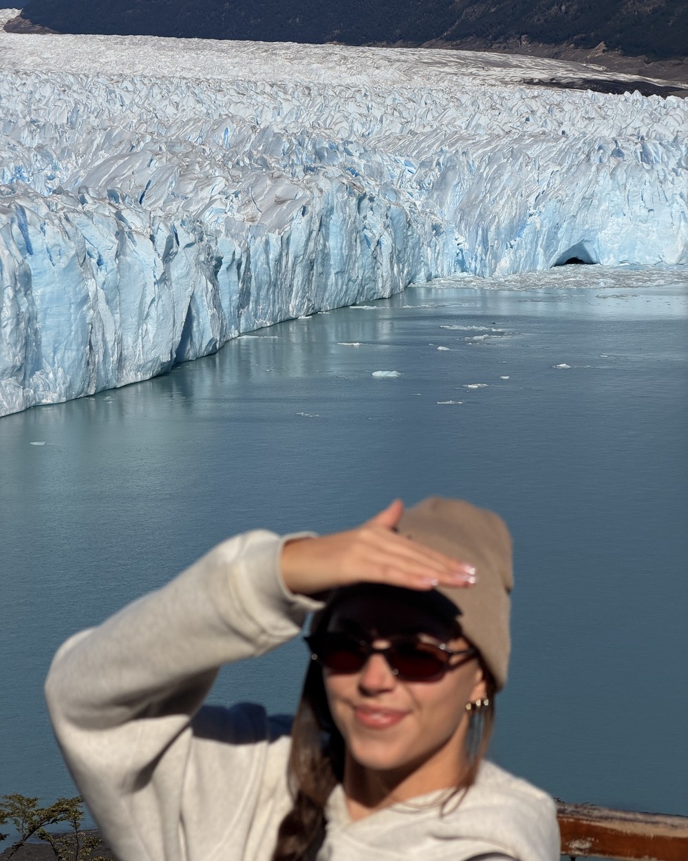 Ellen in a beanie and sunglasses shading her eyes at the walkway railing, with the glacier face and the lake behind her