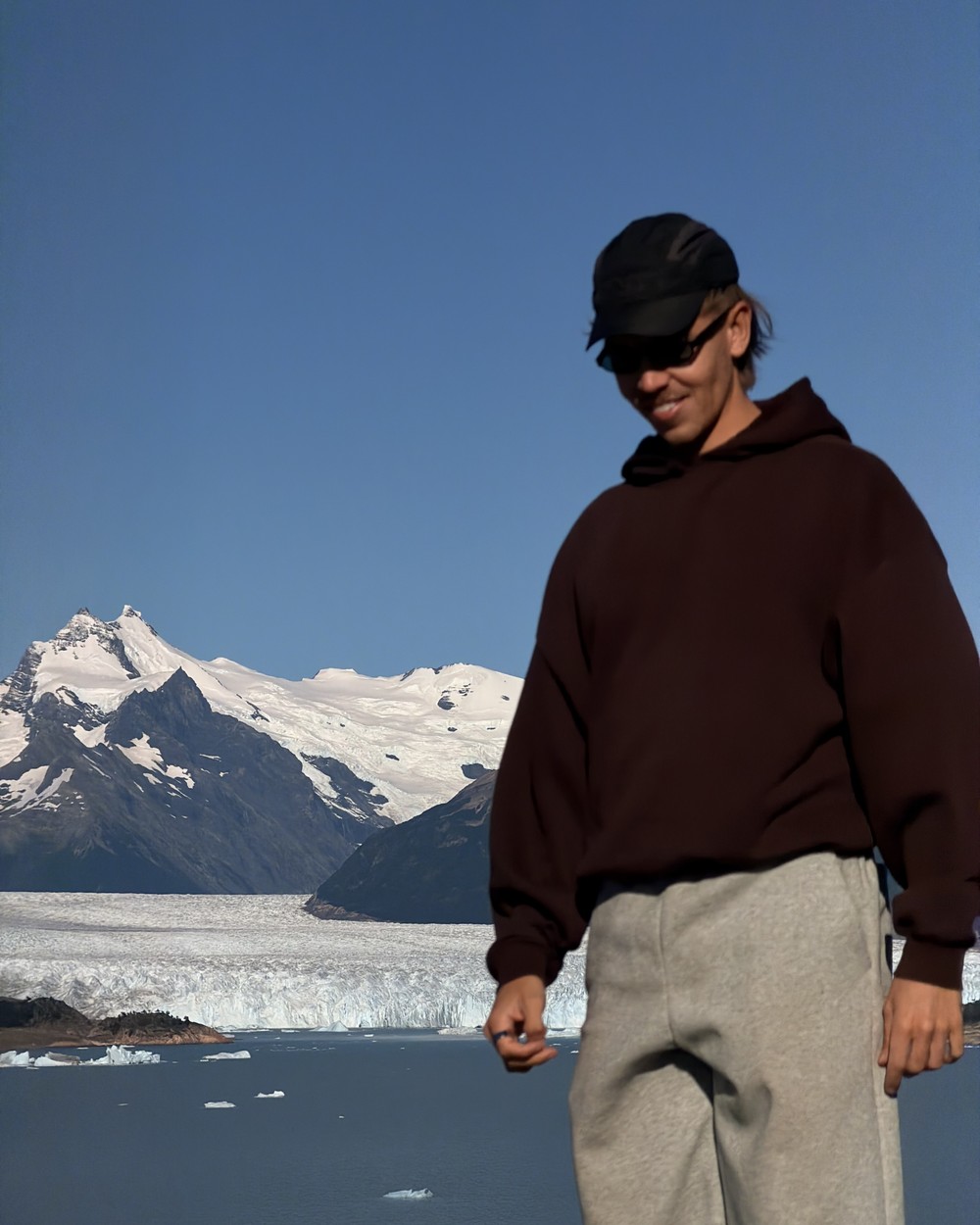 Daniel in a cap and hoodie on the walkway, with the glacier and a snow-covered peak behind him