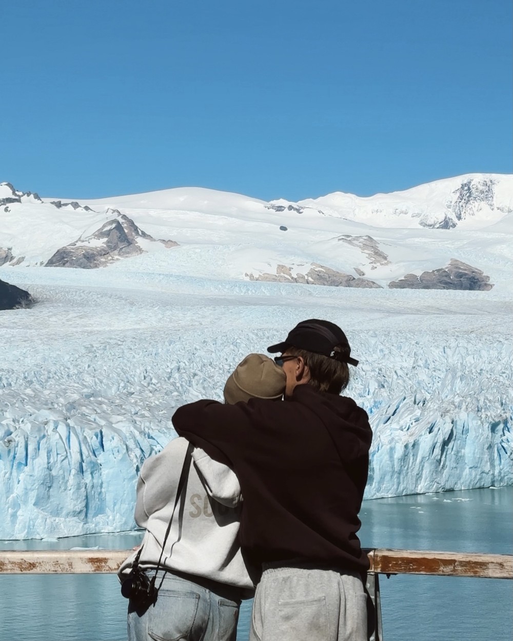 Ellen and Daniel hugging at the walkway railing, with the glacier stretching back to snowy mountains under a blue sky