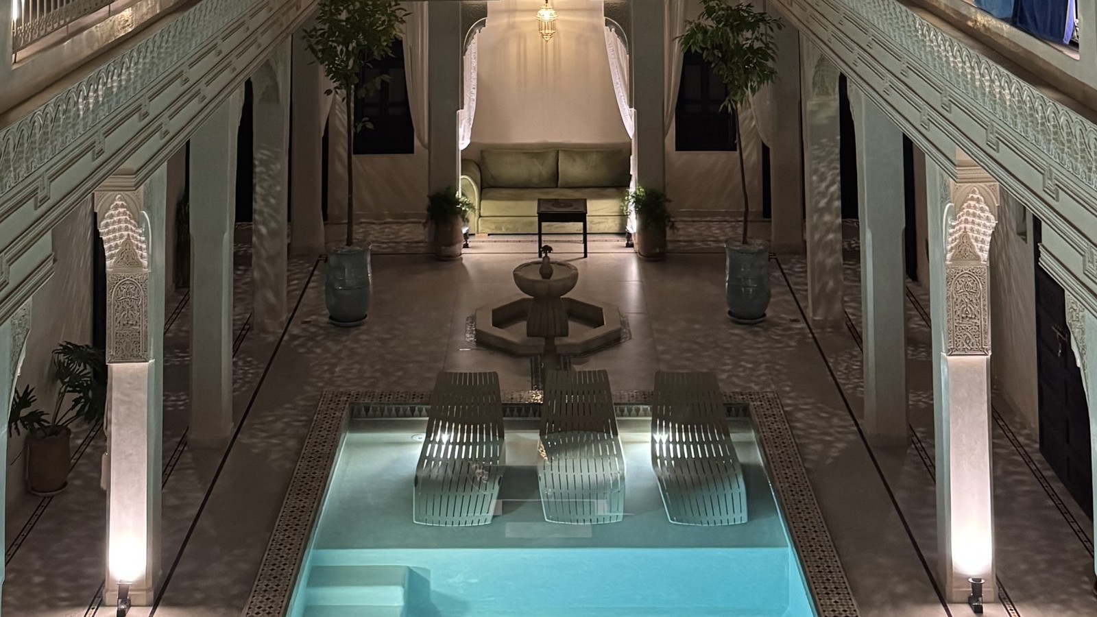 Lit turquoise plunge pool in a riad courtyard at night with arched galleries above in Marrakech, Morocco