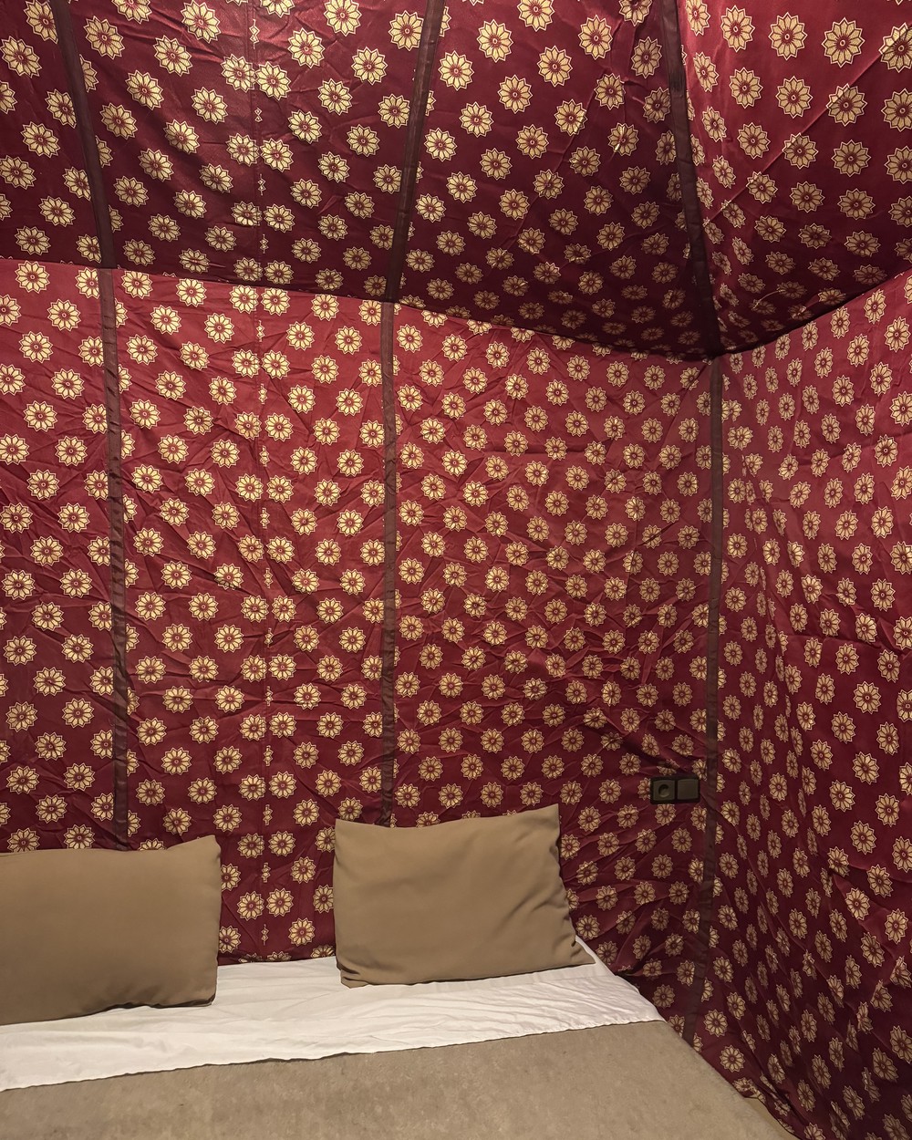Inside a desert camp tent with patterned red fabric walls, a made bed and cushions at Merzouga, Morocco