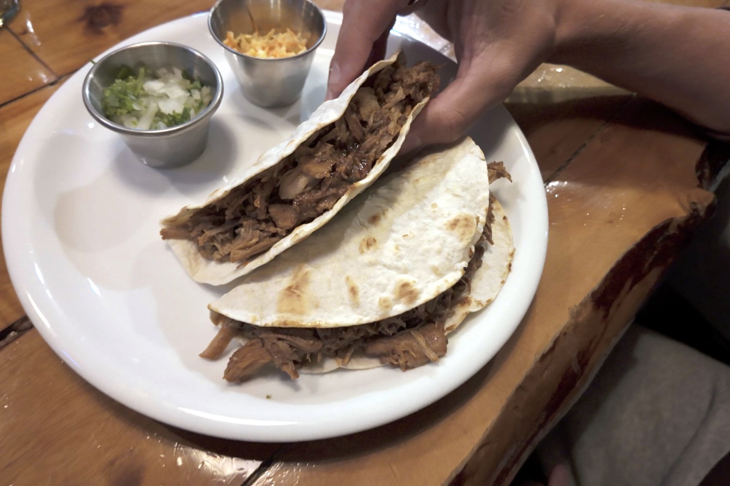Two soft tacos filled with pulled pork on a white plate, with small pots of onion and coriander at Baguales Brewpub
