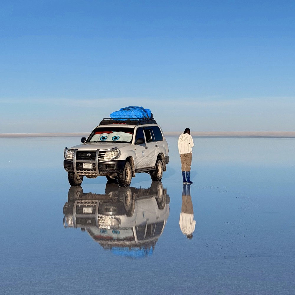 Ellen beside our four-wheel drive reflected on Bolivia's Uyuni salt flats at sunrise