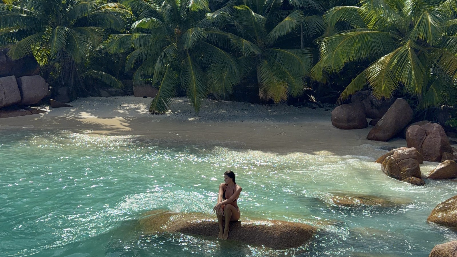 A woman sitting on a granite boulder in shallow turquoise water at the secret beach past Anse Lazio, Praslin, Seychelles