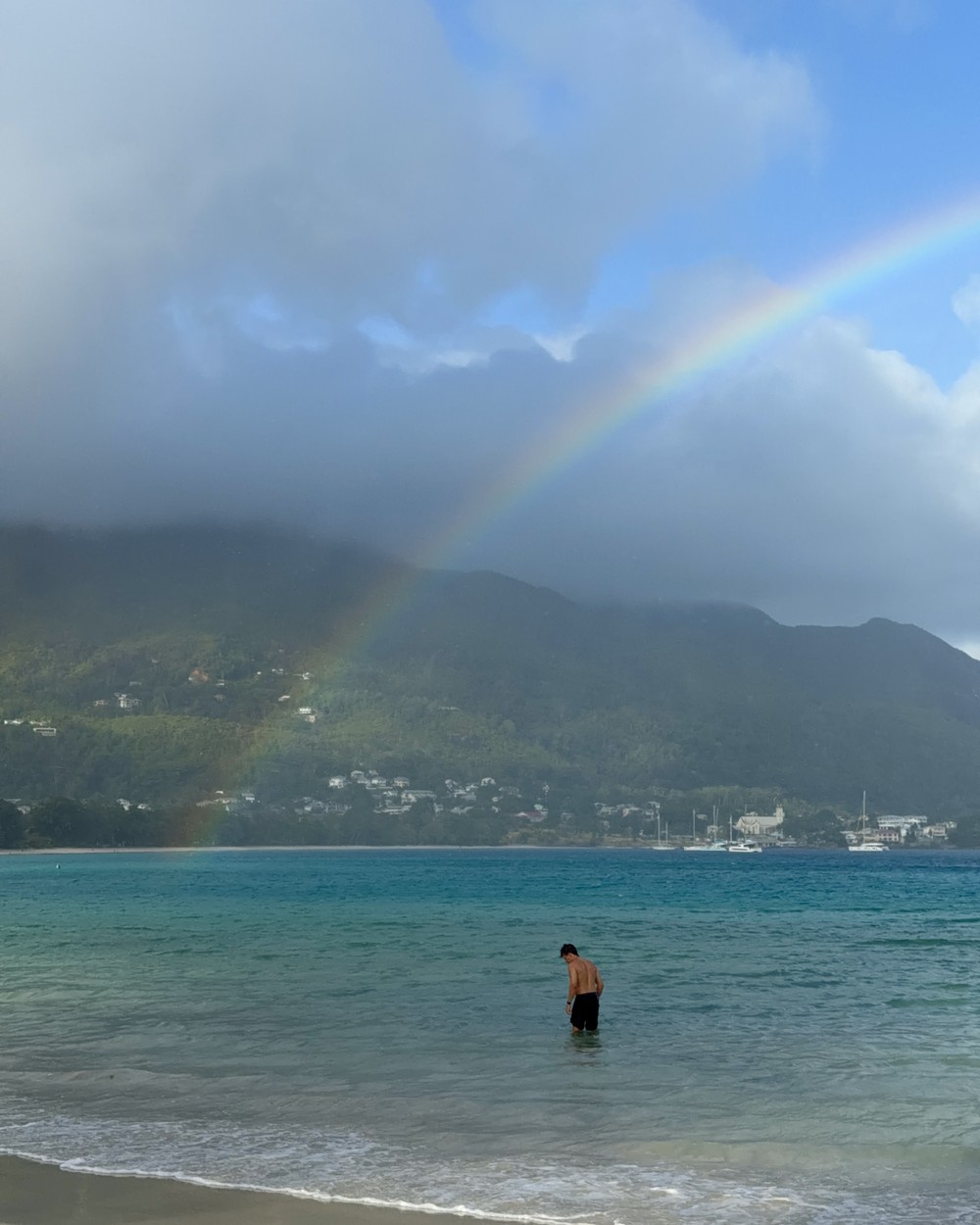 A rainbow coming down onto the flat turquoise water at Beau Vallon on Mahe, Seychelles, with a swimmer standing in the shallows