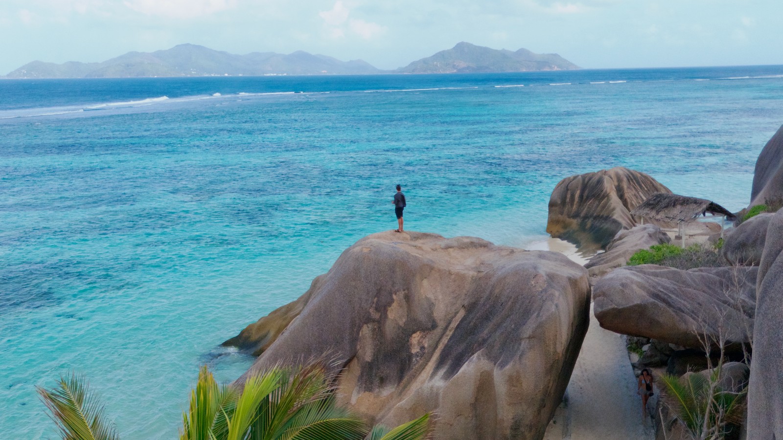 A man standing on a granite boulder above turquoise water at Anse Source d'Argent, La Digue, Seychelles, with an island on the horizon