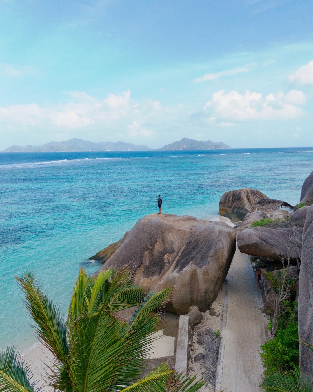 A man standing on a granite boulder above turquoise water at Anse Source d'Argent, La Digue, Seychelles, with an island on the horizon