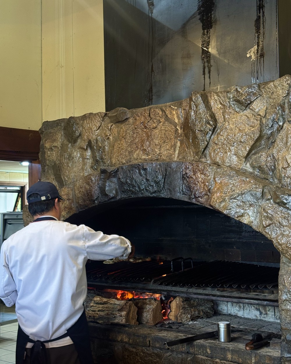 A cook in a white jacket turning meat on a wood-fired grill under a stone arch