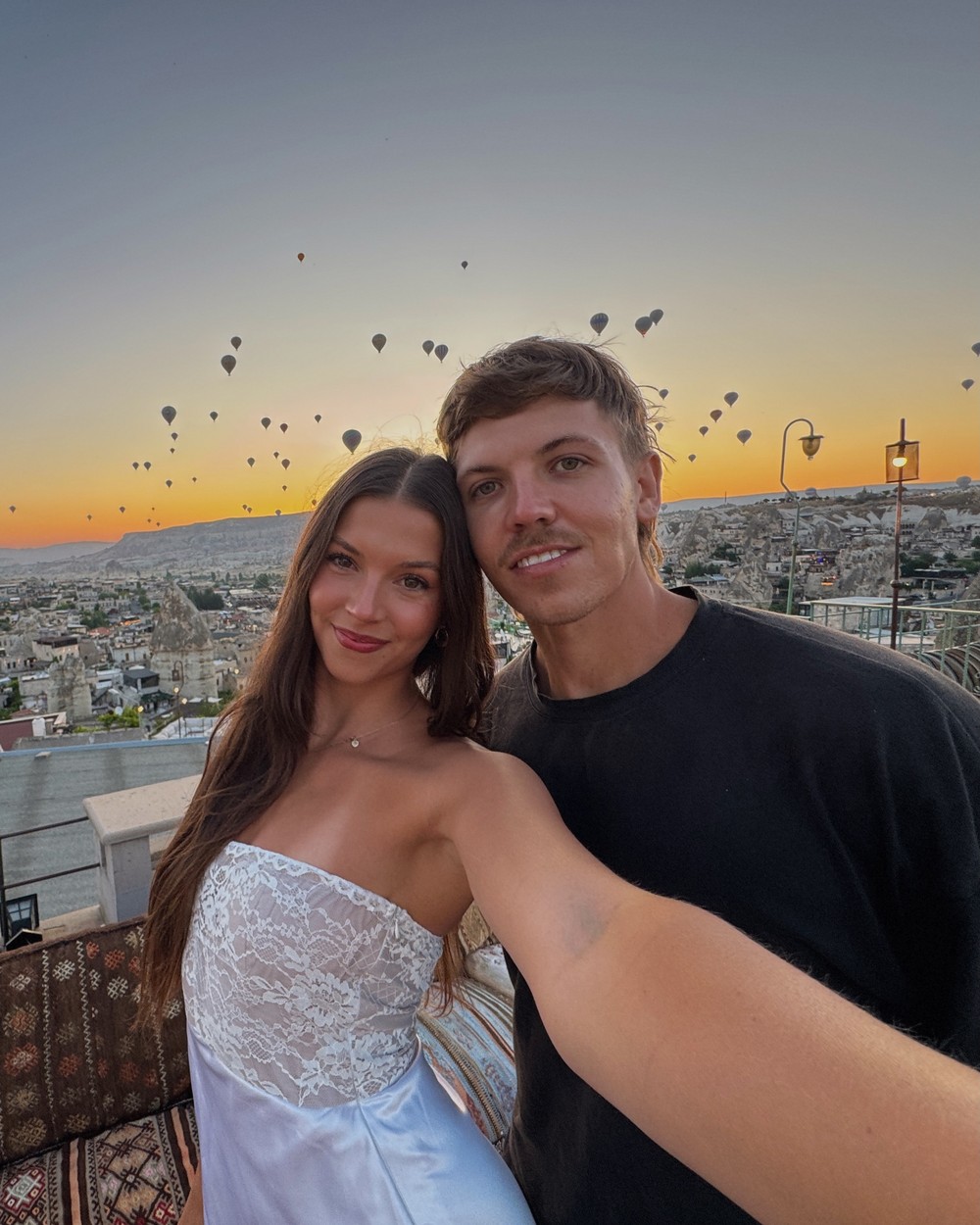 Couple on a hotel rooftop at sunrise with hot air balloons filling the sky behind them