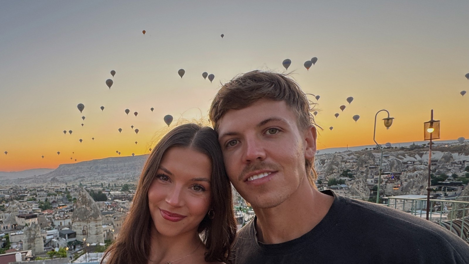 Couple on a cave hotel rooftop at sunrise with hot air balloons rising over Cappadocia